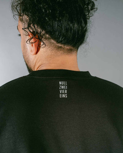 Nahaufnahme der Rückseite eines schwarzen Sweatshirts mit weißem, vertikalem Schriftzug „NULL ZWEI VIER EINS“ im Nackenbereich, getragen von einer Person mit dunklen, lockigen Haaren, die zu einem Dutt gebunden sind. 