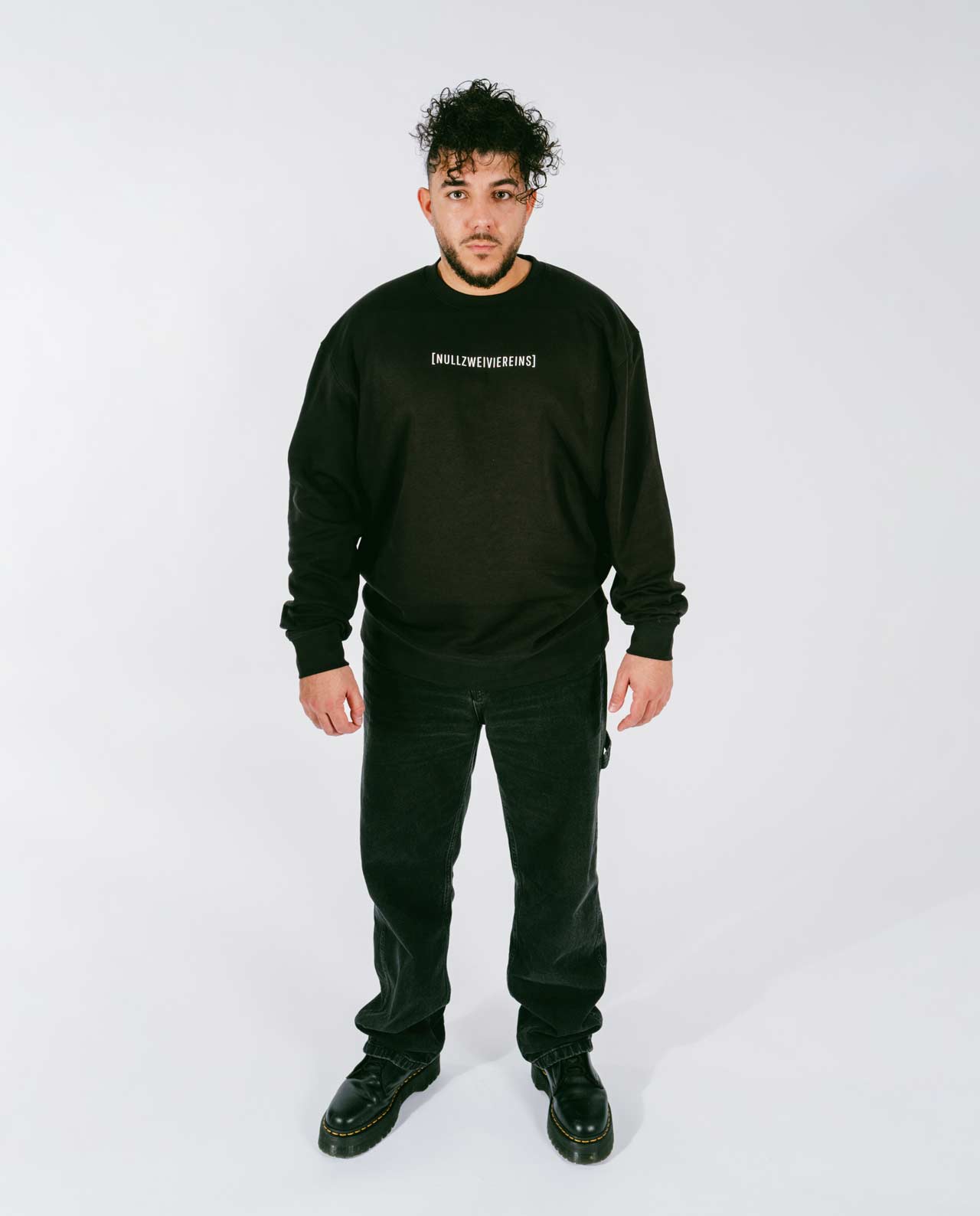 Mann steht frontal vor weißem Hintergrund und trägt einen schwarzen Sweatshirt mit weißem Schriftzug „[NULLZWEIVIEREINS]“ auf der Brust, kombiniert mit schwarzen Jeans und schwarzen Schnürboots.