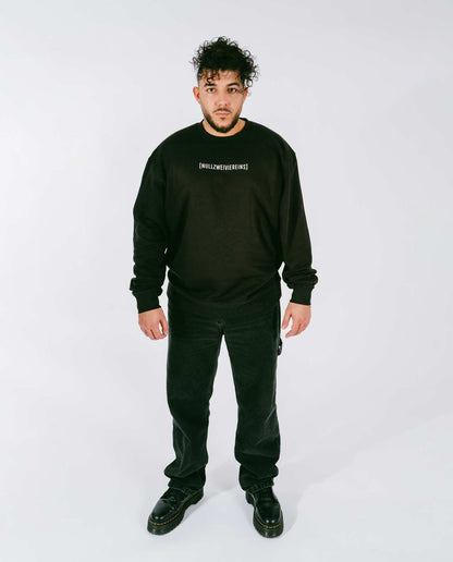 Mann steht frontal vor weißem Hintergrund und trägt einen schwarzen Sweatshirt mit weißem Schriftzug „[NULLZWEIVIEREINS]“ auf der Brust, kombiniert mit schwarzen Jeans und schwarzen Schnürboots.