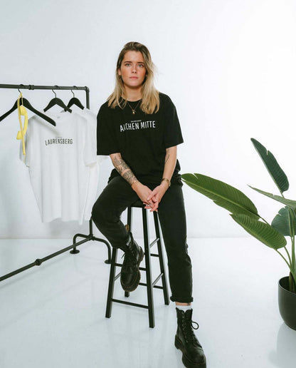 Frau sitzt auf einem hohen schwarzen Hocker und trägt ein schwarzes T-Shirt mit der Aufschrift „AACHEN MITTE“ in weißer, minimalistischer Typografie. Dazu kombiniert sie eine schwarze Jeans und schwarze Boots. Im Hintergrund hängt auf einem Kleiderständer ein weißes T-Shirt mit dem Schriftzug „LAURENSBERG“ sowie ein weiteres weißes Shirt. Rechts im Bild steht eine große grüne Zimmerpflanze.