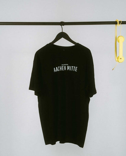 Schwarzes T-Shirt auf einem schwarzen Kleiderbügel, hängend an einer schwarzen Stange. Auf der Brust steht in weißer Schrift „AACHEN MITTE“ mit kleinerem Text darüber „[NULLZWEIVIEREINS]“. Rechts an der Stange hängt ein gelber Telefonhörer mit Spiralkabel.