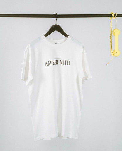 Weißes T-Shirt auf einem schwarzen Kleiderbügel, hängend an einer schwarzen Stange. Auf der Brust steht in schwarzer Schrift „AACHEN MITTE“ mit kleinerem Text darüber „[NULLZWEIVIEREINS]“. Rechts an der Stange hängt ein gelber Telefonhörer mit Spiralkabel.