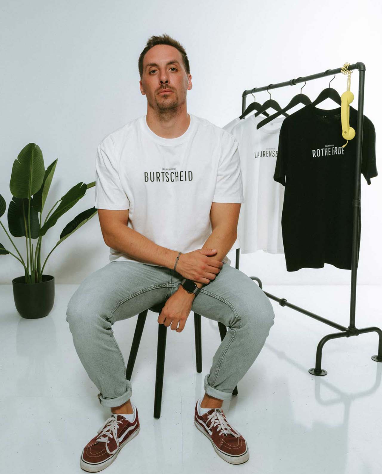 Mann sitzt auf einem Hocker in einem weißen T-Shirt mit der Aufschrift „BURTSCHEID“ in minimalistischer schwarzer Typografie. Neben ihm steht ein Kleiderständer mit weiteren T-Shirts in Weiß („LAURENSBERG“) und Schwarz („ROTHE ERDE“). Im Hintergrund steht eine grüne Zimmerpflanze, auf dem Kleiderständer hängt ein gelber Telefonhörer als Dekoration.
