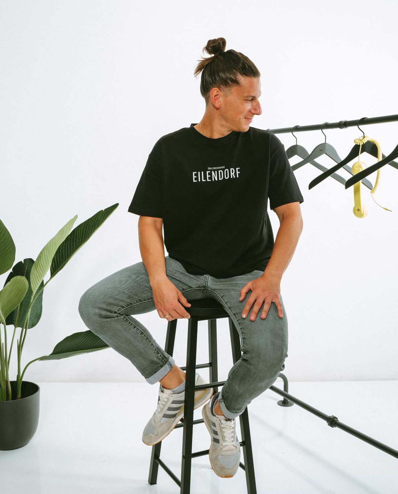 Mann sitzt seitlich auf einem hohen schwarzen Hocker und trägt ein schwarzes T-Shirt mit der Aufschrift „EILENDORF“ in klarer, weißer Typografie. Er schaut nach rechts und lächelt leicht. Neben ihm steht ein Kleiderständer mit leeren schwarzen Kleiderbügeln und einem gelben Telefonhörer als Dekoration. Im Hintergrund ist eine grüne Zimmerpflanze zu sehen.
