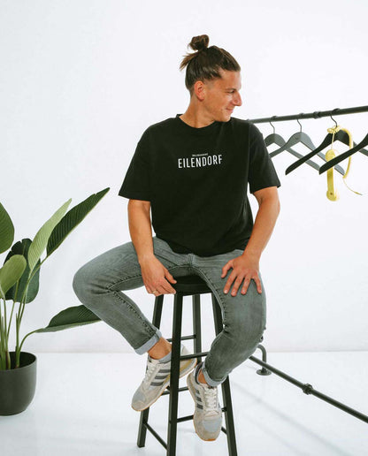Mann sitzt seitlich auf einem hohen schwarzen Hocker und trägt ein schwarzes T-Shirt mit der Aufschrift „EILENDORF“ in klarer, weißer Typografie. Er schaut nach rechts und lächelt leicht. Neben ihm steht ein Kleiderständer mit leeren schwarzen Kleiderbügeln und einem gelben Telefonhörer als Dekoration. Im Hintergrund ist eine grüne Zimmerpflanze zu sehen.