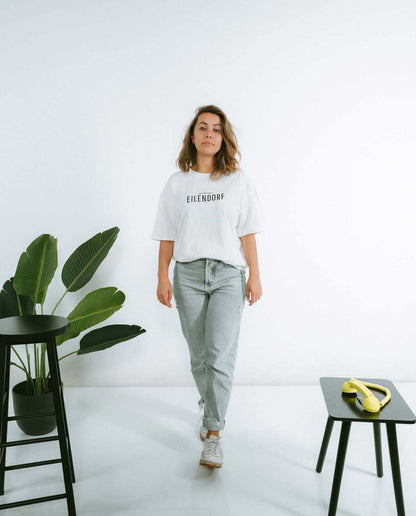 Frau geht selbstbewusst auf die Kamera zu und trägt ein weißes T-Shirt mit der Aufschrift „EILENDORF“ in schwarzer, klarer Typografie. Sie kombiniert es mit einer hellgrauen Jeans und weißen Sneakers. Links steht ein schwarzer Hocker neben einer grünen Zimmerpflanze, rechts ein kleiner schwarzer Tisch mit einem gelben Telefonhörer als Deko.