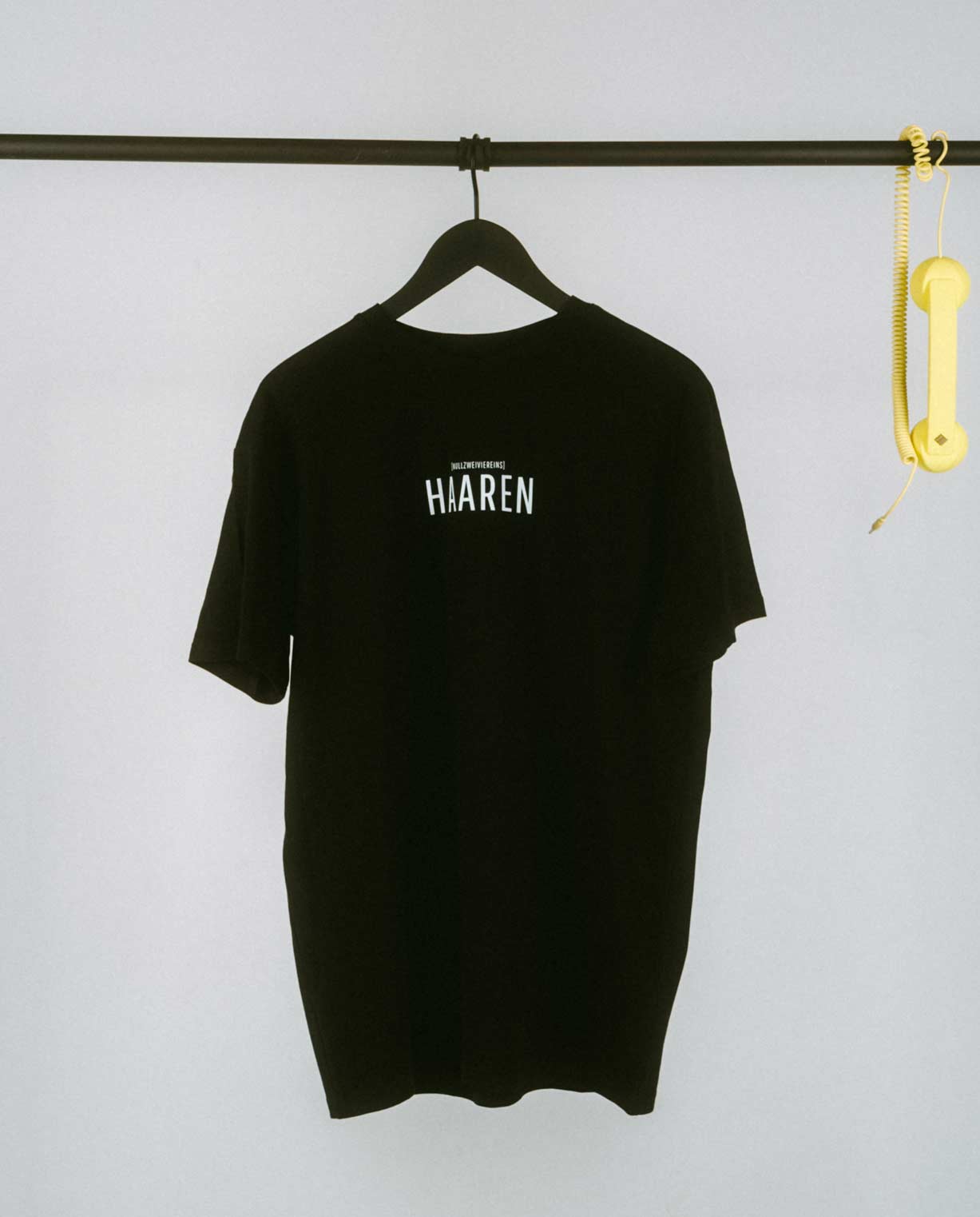 Minimalistisches schwarzes T-Shirt auf einem schwarzen Kleiderbügel, aufgehängt an einer schlichten weißen Kleiderstange. Auf der Brust ist mittig in großen schwarzen Großbuchstaben der Stadtteilname „HAAREN“ aufgedruckt, darüber in kleinerer Schrift der Zusatz „[NULLZWEIVIEREINS]“. Rechts an der Stange hängt als verspieltes Detail ein gelber Retro-Telefonhörer mit Spiralkabel, der einen farbigen Akzent zum cleanen und hellen Hintergrund setzt.