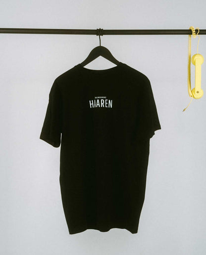 Minimalistisches schwarzes T-Shirt auf einem schwarzen Kleiderbügel, aufgehängt an einer schlichten weißen Kleiderstange. Auf der Brust ist mittig in großen schwarzen Großbuchstaben der Stadtteilname „HAAREN“ aufgedruckt, darüber in kleinerer Schrift der Zusatz „[NULLZWEIVIEREINS]“. Rechts an der Stange hängt als verspieltes Detail ein gelber Retro-Telefonhörer mit Spiralkabel, der einen farbigen Akzent zum cleanen und hellen Hintergrund setzt.