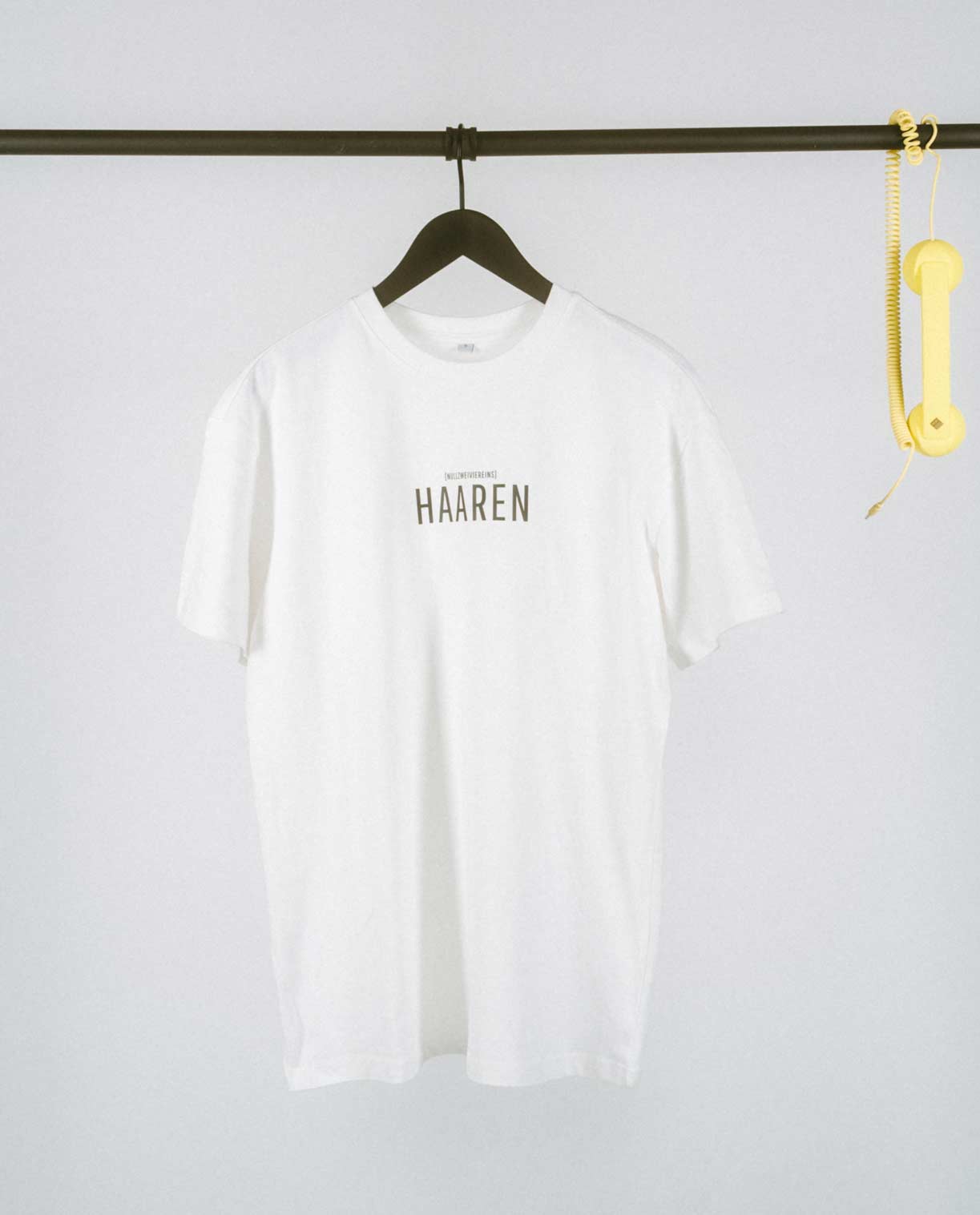Minimalistisches weißes T-Shirt auf einem schwarzen Kleiderbügel, aufgehängt an einer schlichten schwarzen Kleiderstange. Auf der Brust ist mittig in großen schwarzen Großbuchstaben der Stadtteilname „HAAREN“ aufgedruckt, darüber in kleinerer Schrift der Zusatz „[NULLZWEIVIEREINS]“. Rechts an der Stange hängt als verspieltes Detail ein gelber Retro-Telefonhörer mit Spiralkabel, der einen farbigen Akzent zum cleanen und hellen Hintergrund setzt.