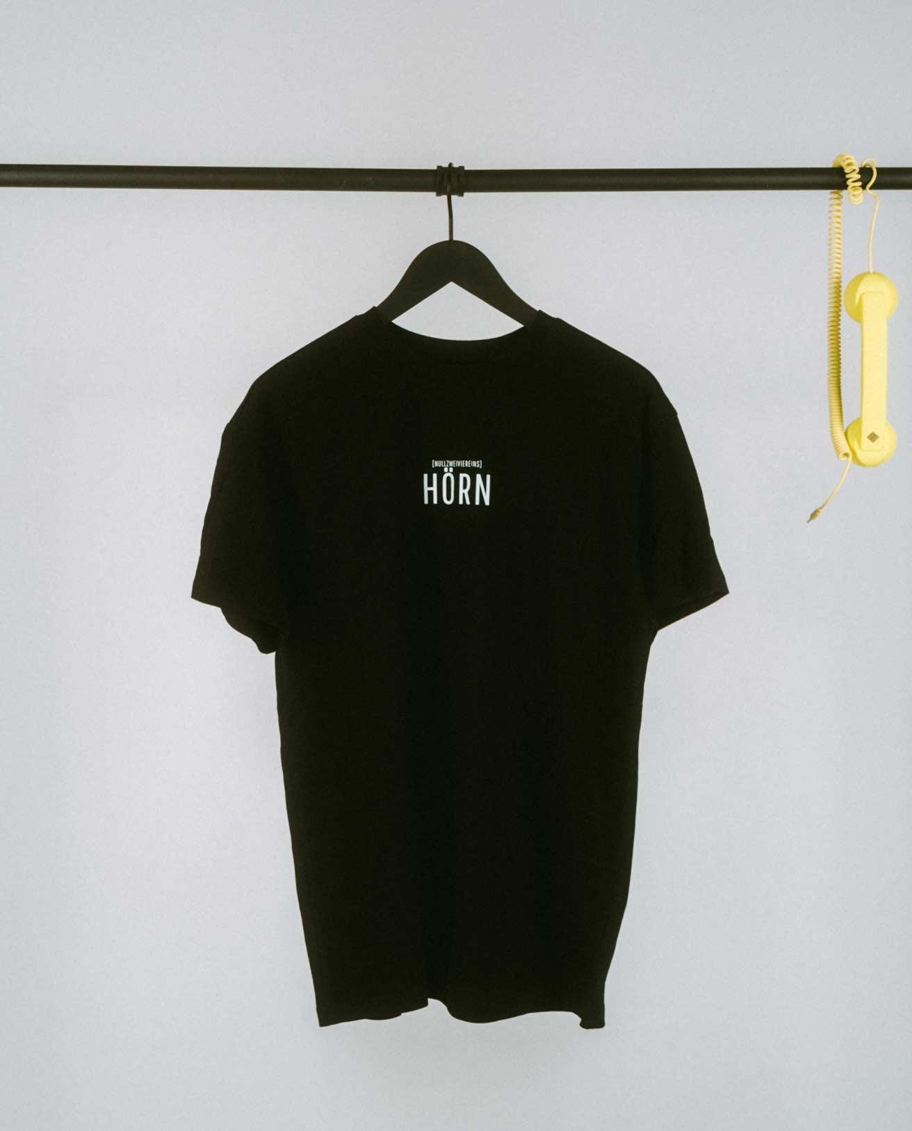 Schlichtes schwarzes T-Shirt auf einem schwarzen Kleiderbügel, präsentiert an einer minimalistischen schwarzen Kleiderstange. Auf der Vorderseite steht mittig in weißen Großbuchstaben „HÖRN“, darüber in kleinerer Schrift der Zusatz „[NULLZWEIVIEREINS]“. Rechts an der Stange hängt als verspieltes Detail ein gelber Telefonhörer mit Spiralkabel. Der helle Hintergrund verstärkt den cleanen und modernen Look des Shirts.
