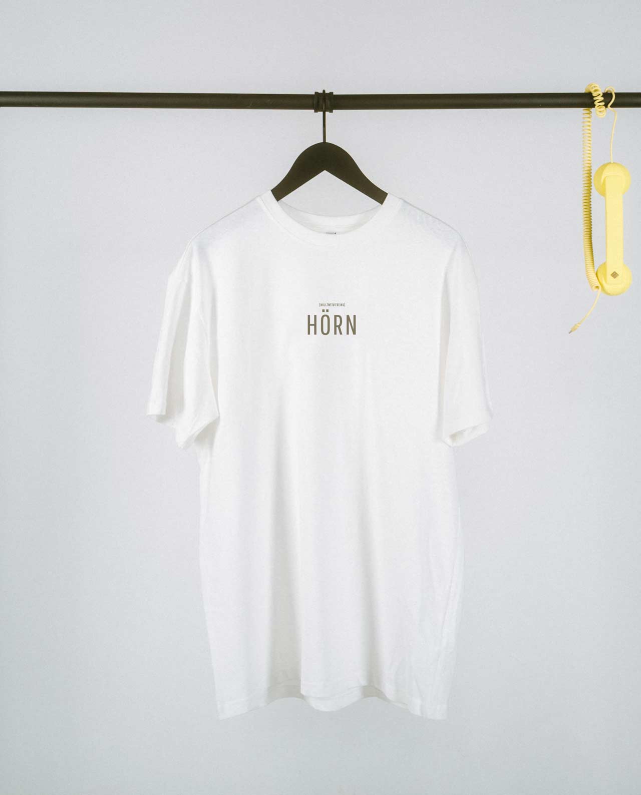 Schlichtes weißes T-Shirt auf einem schwarzen Kleiderbügel, präsentiert an einer minimalistischen schwarzen Kleiderstange. Auf der Vorderseite steht mittig in schwarzen Großbuchstaben „HÖRN“, darüber in kleinerer Schrift der Zusatz „[NULLZWEIVIEREINS]“. Rechts an der Stange hängt als verspieltes Detail ein gelber Telefonhörer mit Spiralkabel. Der helle Hintergrund verstärkt den cleanen und modernen Look des Shirts.