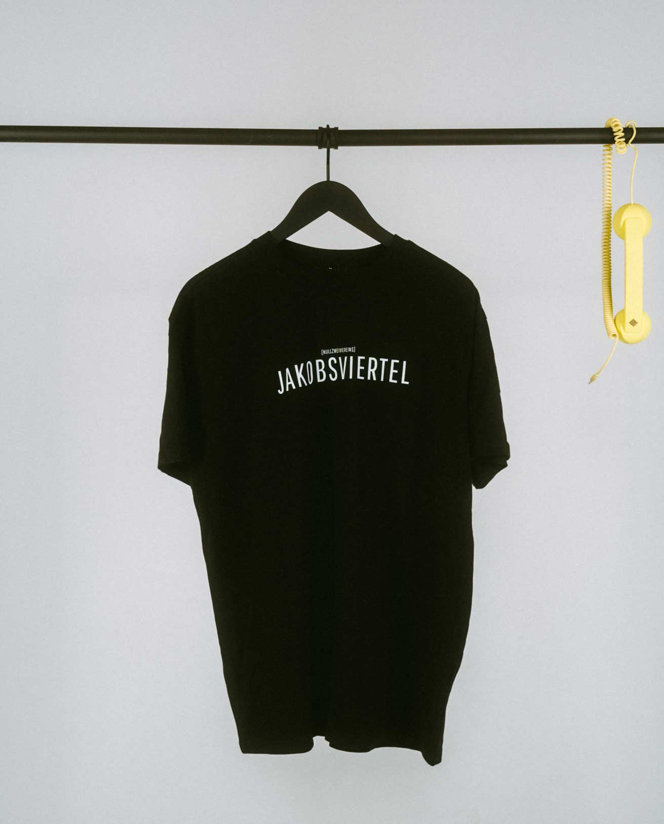 Schwarzes T-Shirt auf einem schwarzen Bügel, aufgehängt an einer schlichten schwarzen Kleiderstange. Zentral auf der Vorderseite steht in weißen Großbuchstaben „JAKOBSVIERTEL“, darüber in kleinerer Schrift der Zusatz „[NULLZWEIVIEREINS]“. Rechts an der Stange hängt ein gelber Telefonhörer mit Spiralkabel als auffälliges Deko-Element.