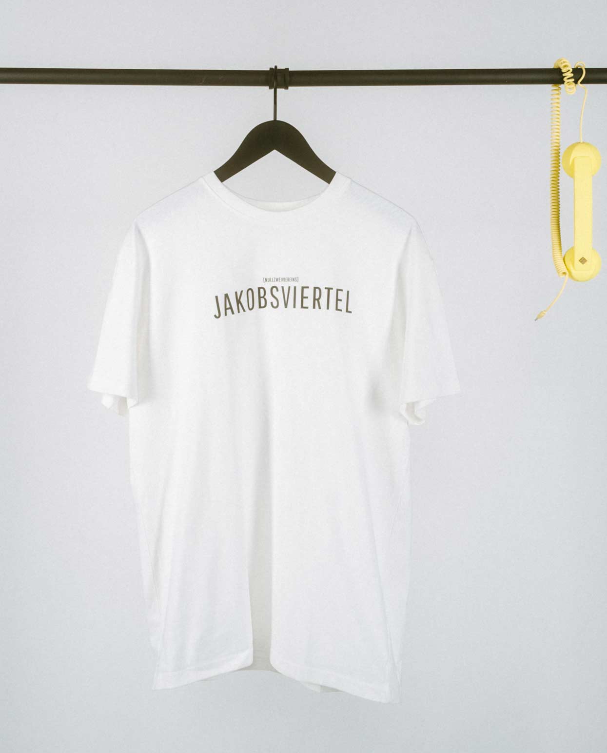 Weißes T-Shirt auf einem schwarzen Bügel, aufgehängt an einer schlichten schwarzen Kleiderstange. Zentral auf der Vorderseite steht in schwarzen Großbuchstaben „JAKOBSVIERTEL“, darüber in kleinerer Schrift der Zusatz „[NULLZWEIVIEREINS]“. Rechts an der Stange hängt ein gelber Telefonhörer mit Spiralkabel als auffälliges Deko-Element.