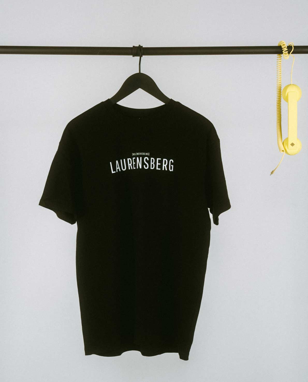 Schlichtes, schwarzes T-Shirt auf einem schwarzen Kleiderbügel, hängend an einer schwarzen Metallstange. Auf der Vorderseite mittig der Aufdruck „LAURENSBERG“ in klaren, weißen Großbuchstaben, darüber in kleinerer Schrift der Zusatz „[NULLZWEIVIEREINS]“. Rechts an der Stange hängt dekorativ ein gelber Telefonhörer mit Spiralkabel.