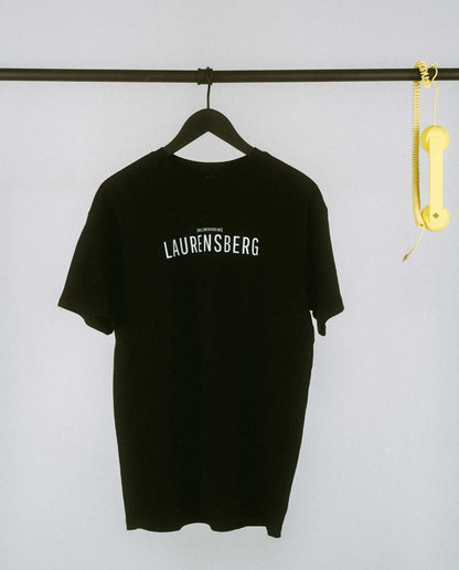 Schlichtes, schwarzes T-Shirt auf einem schwarzen Kleiderbügel, hängend an einer schwarzen Metallstange. Auf der Vorderseite mittig der Aufdruck „LAURENSBERG“ in klaren, weißen Großbuchstaben, darüber in kleinerer Schrift der Zusatz „[NULLZWEIVIEREINS]“. Rechts an der Stange hängt dekorativ ein gelber Telefonhörer mit Spiralkabel.