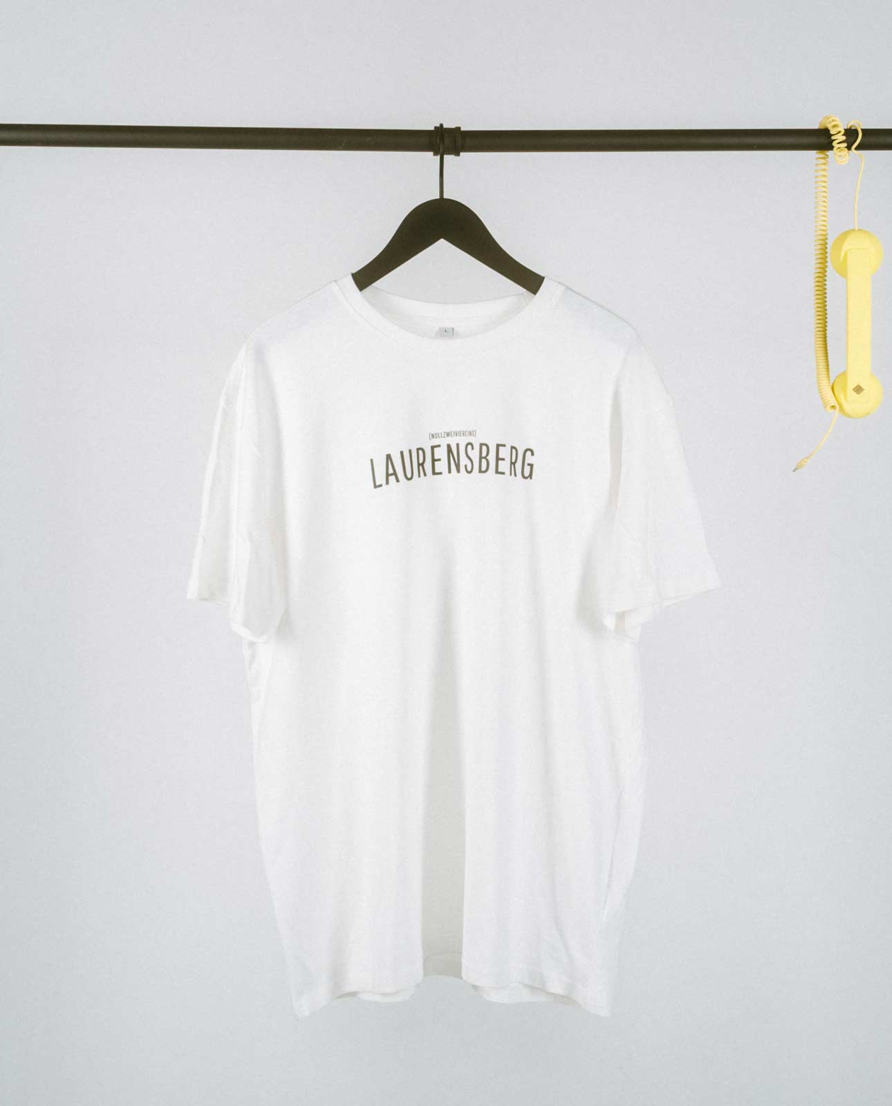 Schlichtes, weißes T-Shirt auf einem schwarzen Kleiderbügel, hängend an einer schwarzen Metallstange. Auf der Vorderseite mittig der Aufdruck „LAURENSBERG“ in klaren, schwarzen Großbuchstaben, darüber in kleinerer Schrift der Zusatz „[NULLZWEIVIEREINS]“. Rechts an der Stange hängt dekorativ ein gelber Telefonhörer mit Spiralkabel.