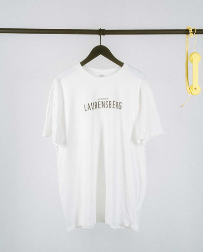 Schlichtes, weißes T-Shirt auf einem schwarzen Kleiderbügel, hängend an einer schwarzen Metallstange. Auf der Vorderseite mittig der Aufdruck „LAURENSBERG“ in klaren, schwarzen Großbuchstaben, darüber in kleinerer Schrift der Zusatz „[NULLZWEIVIEREINS]“. Rechts an der Stange hängt dekorativ ein gelber Telefonhörer mit Spiralkabel.