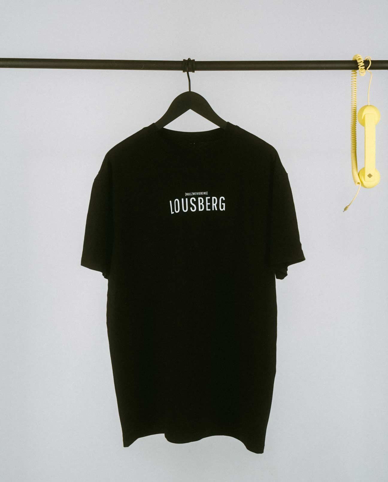 Schwarzes, locker geschnittenes T-Shirt, das auf einem schwarzen Kleiderbügel an einer schwarzen Metallstange hängt. Auf der Brust steht in schlichter, weißer Großbuchstaben-Typografie „LOUSBERG“, darüber in kleinerer Schrift „[NULLZWEIVIEREINS]“. Rechts an der Stange hängt als dekoratives Element ein gelber Telefonhörer mit Spiralkabel. Der Hintergrund ist schlicht und hell, wodurch das minimalistische Design des T-Shirts im Vordergrund steht.