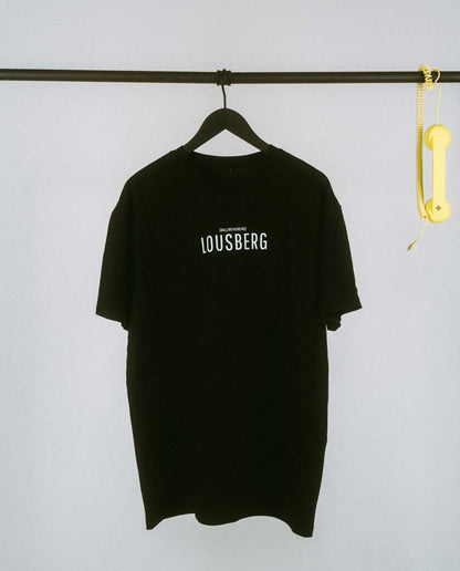 Schwarzes, locker geschnittenes T-Shirt, das auf einem schwarzen Kleiderbügel an einer schwarzen Metallstange hängt. Auf der Brust steht in schlichter, weißer Großbuchstaben-Typografie „LOUSBERG“, darüber in kleinerer Schrift „[NULLZWEIVIEREINS]“. Rechts an der Stange hängt als dekoratives Element ein gelber Telefonhörer mit Spiralkabel. Der Hintergrund ist schlicht und hell, wodurch das minimalistische Design des T-Shirts im Vordergrund steht.
