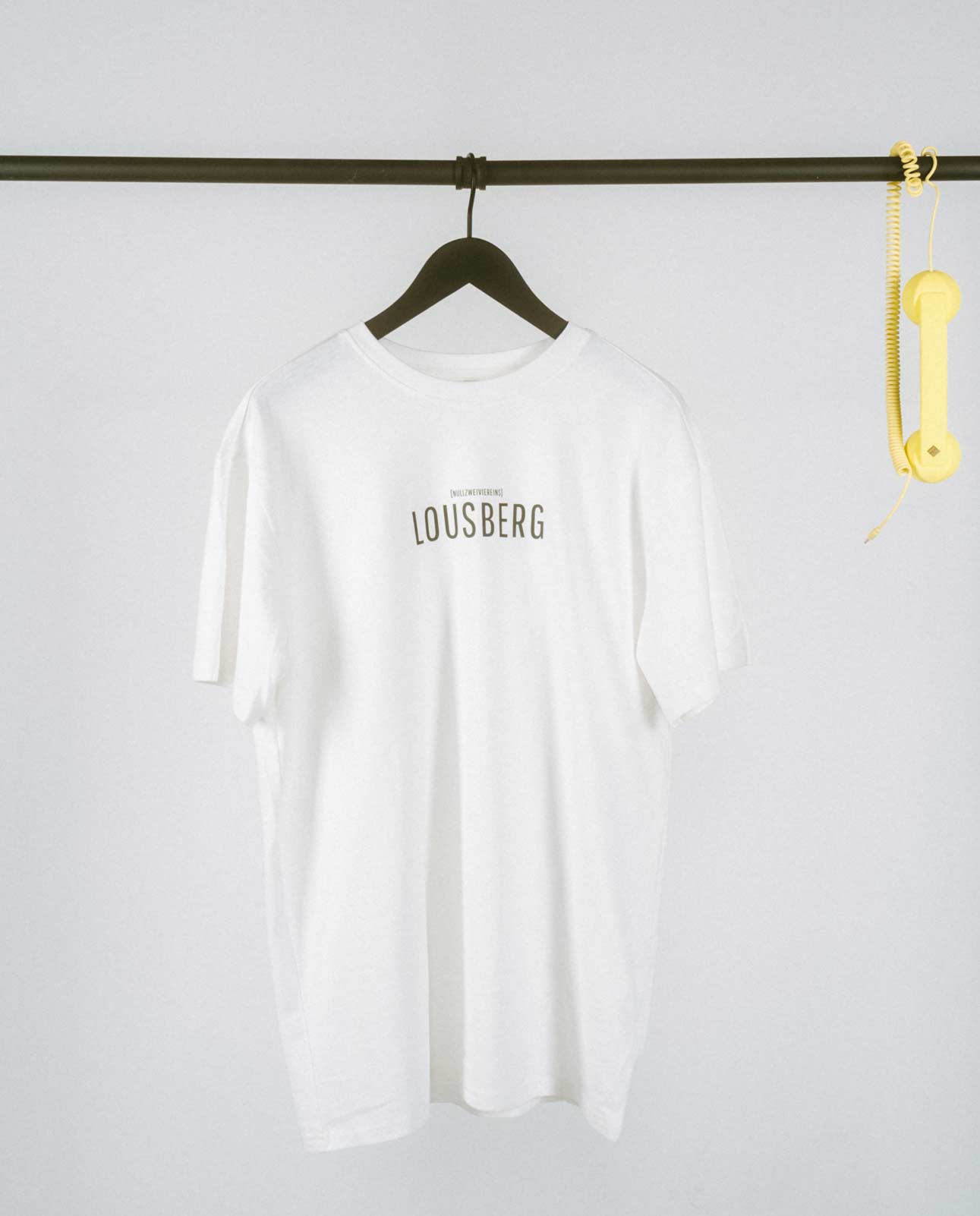 Weißes, locker geschnittenes T-Shirt, das auf einem schwarzen Kleiderbügel an einer schwarzen Metallstange hängt. Auf der Brust steht in schlichter, schwarzer Großbuchstaben-Typografie „LOUSBERG“, darüber in kleinerer Schrift „[NULLZWEIVIEREINS]“. Rechts an der Stange hängt als dekoratives Element ein gelber Telefonhörer mit Spiralkabel. Der Hintergrund ist schlicht und hell, wodurch das minimalistische Design des T-Shirts im Vordergrund steht.
