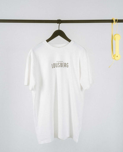 Weißes, locker geschnittenes T-Shirt, das auf einem schwarzen Kleiderbügel an einer schwarzen Metallstange hängt. Auf der Brust steht in schlichter, schwarzer Großbuchstaben-Typografie „LOUSBERG“, darüber in kleinerer Schrift „[NULLZWEIVIEREINS]“. Rechts an der Stange hängt als dekoratives Element ein gelber Telefonhörer mit Spiralkabel. Der Hintergrund ist schlicht und hell, wodurch das minimalistische Design des T-Shirts im Vordergrund steht.