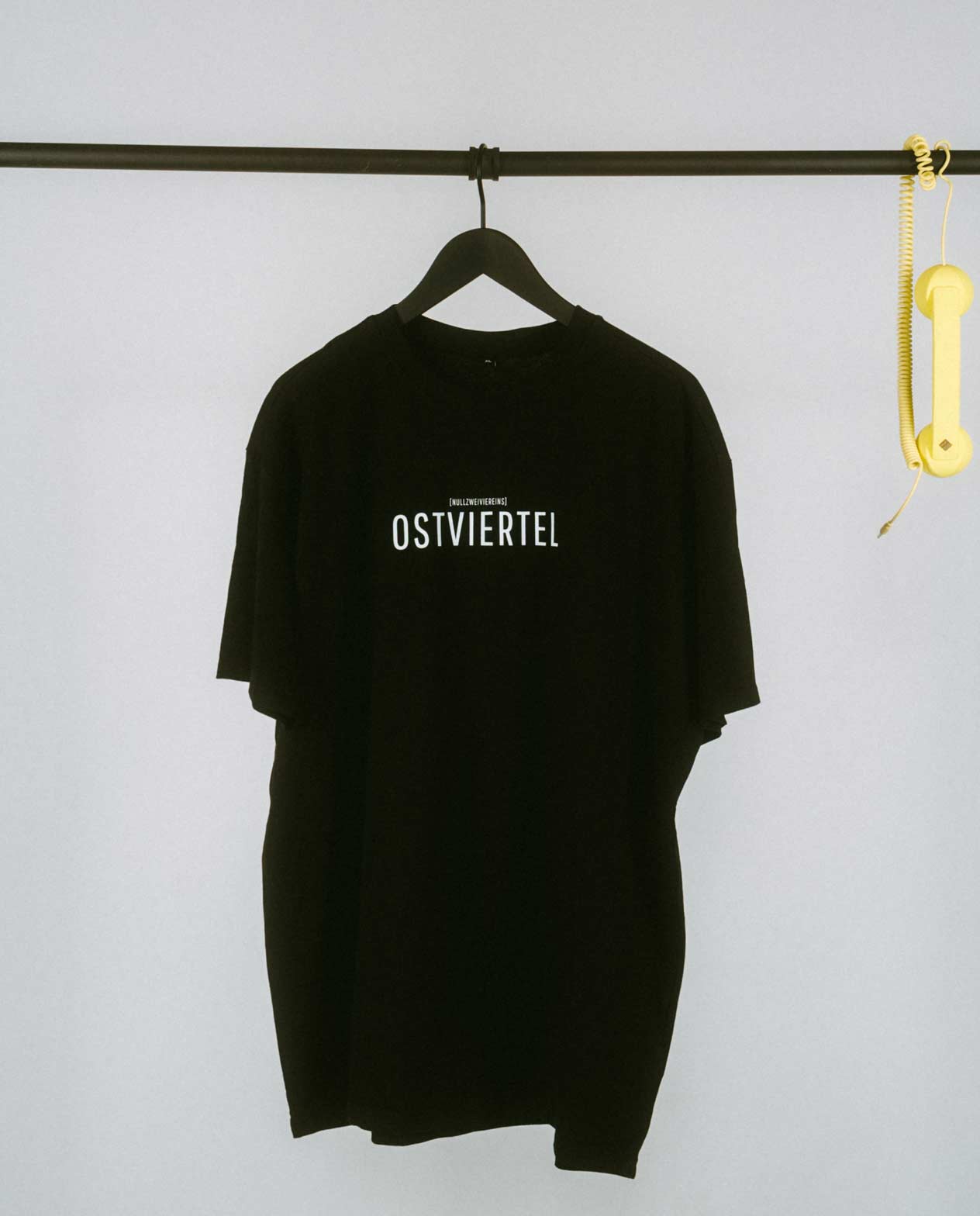 Weißes T-Shirt auf einem schwarzen Kleiderbügel, hängend an einer schwarzen Stange. Auf der Brust steht in schwarzer Schrift „OSTVIERTEL“ mit kleinerem Text darüber „[NULLZWEIVIEREINS]“. Rechts an der Stange hängt ein gelber Telefonhörer mit Spiralkabel.