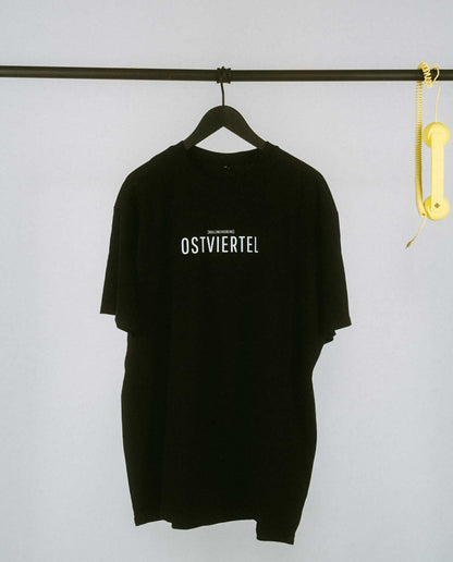 Weißes T-Shirt auf einem schwarzen Kleiderbügel, hängend an einer schwarzen Stange. Auf der Brust steht in schwarzer Schrift „OSTVIERTEL“ mit kleinerem Text darüber „[NULLZWEIVIEREINS]“. Rechts an der Stange hängt ein gelber Telefonhörer mit Spiralkabel.