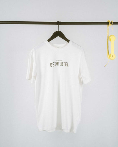 Weißes T-Shirt auf einem schwarzen Kleiderbügel, hängend an einer schwarzen Stange. Auf der Brust steht in schwarzer Schrift „OSTVIERTEL“ mit kleinerem Text darüber „[NULLZWEIVIEREINS]“. Rechts an der Stange hängt ein gelber Telefonhörer mit Spiralkabel.