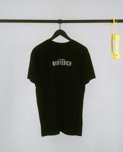 Schwarzes T-Shirt auf einem schwarzen Kleiderbügel, hängend an einer schwarzen Stange. Auf der Brust steht in schwarzer Schrift „RICHTERICH“ mit kleinerem Text darüber „[NULLZWEIVIEREINS]“. Rechts an der Stange hängt ein gelber Telefonhörer mit Spiralkabel.