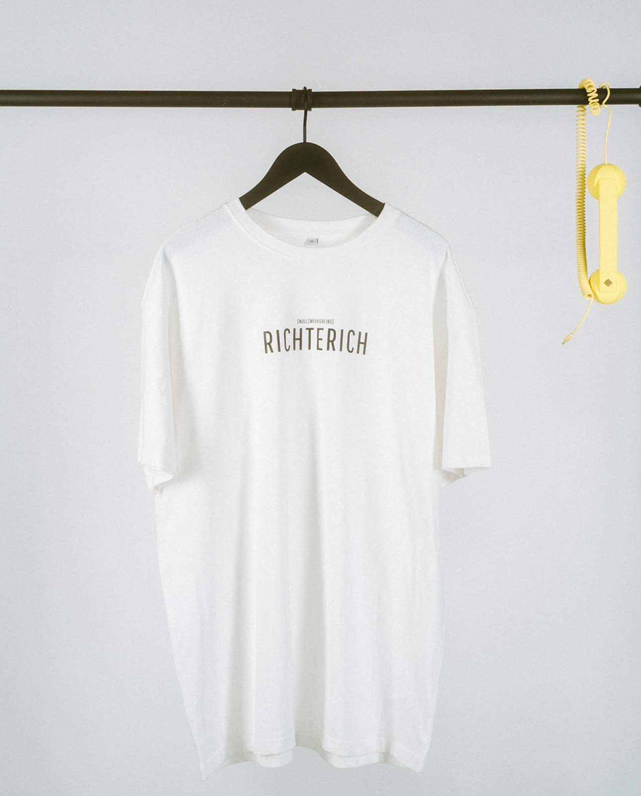 Weißes T-Shirt auf einem schwarzen Kleiderbügel, hängend an einer schwarzen Stange. Auf der Brust steht in schwarzer Schrift „RICHTERICH“ mit kleinerem Text darüber „[NULLZWEIVIEREINS]“. Rechts an der Stange hängt ein gelber Telefonhörer mit Spiralkabel.