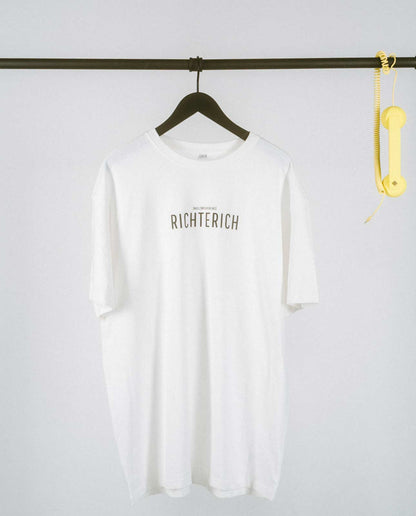 Weißes T-Shirt auf einem schwarzen Kleiderbügel, hängend an einer schwarzen Stange. Auf der Brust steht in schwarzer Schrift „RICHTERICH“ mit kleinerem Text darüber „[NULLZWEIVIEREINS]“. Rechts an der Stange hängt ein gelber Telefonhörer mit Spiralkabel.