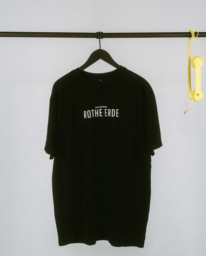 Schwarzes T-Shirt auf einem Kleiderbügel, hängend an einer schwarzen Stange. Auf der Brust in weißer Schrift der Aufdruck „ROTHE ERDE“ mit kleinerem Text darüber „[NULLZWEIVIEREINS]“. Rechts an der Stange hängt ein gelber Telefonhörer mit Spiralkabel.