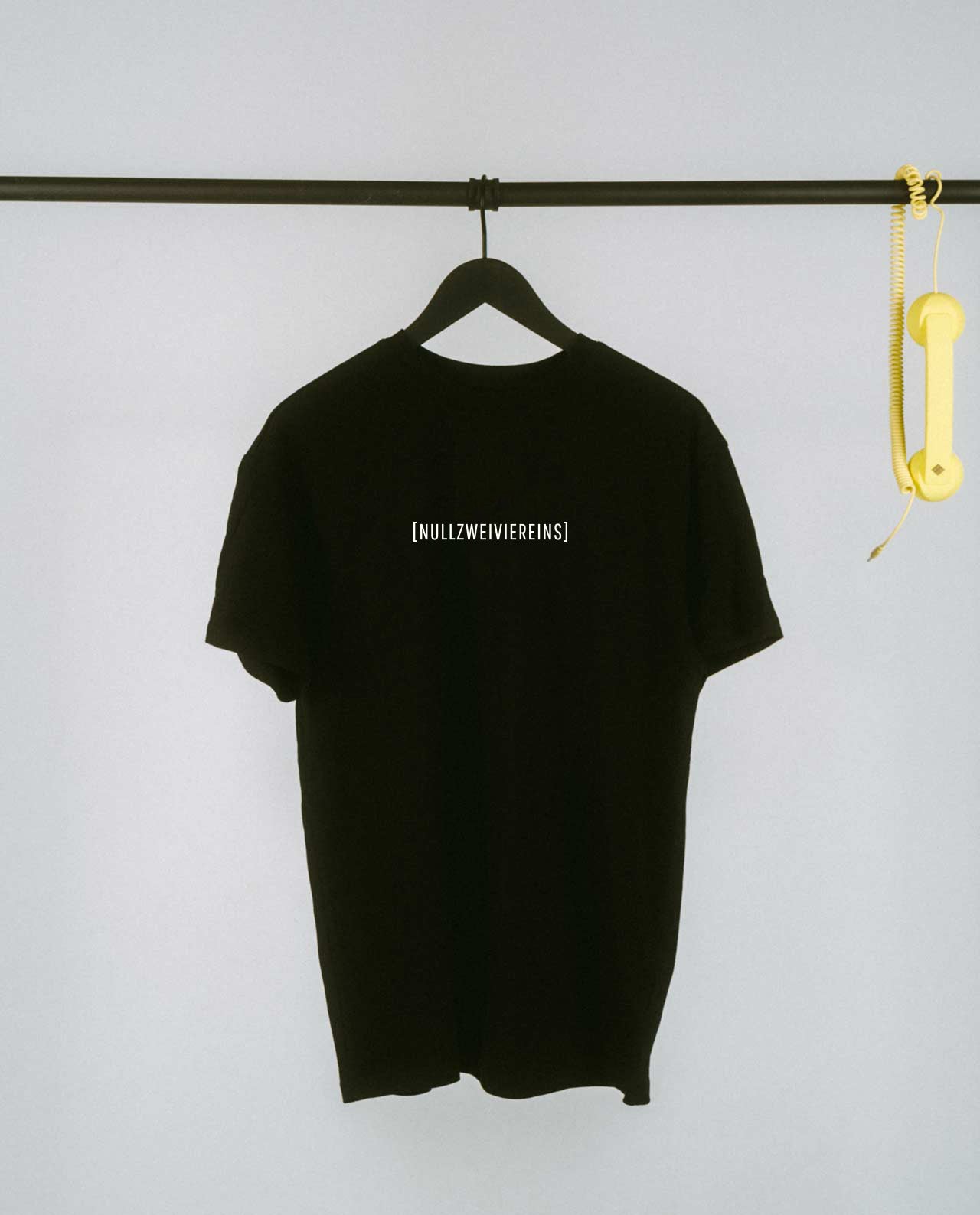 Schwarzes, locker geschnittenes T-Shirt, das auf einem schwarzen Kleiderbügel an einer schwarzen Metallstange hängt. Mittig auf der Brust steht in schlichter schwarzer Schrift „[NULLZWEIVIEREINS]“. Rechts an der Stange baumelt ein gelber Telefonhörer mit Spiralkabel als dekoratives Element. Der Hintergrund ist schlicht und hell, wodurch das T-Shirt im Vordergrund klar zur Geltung kommt.