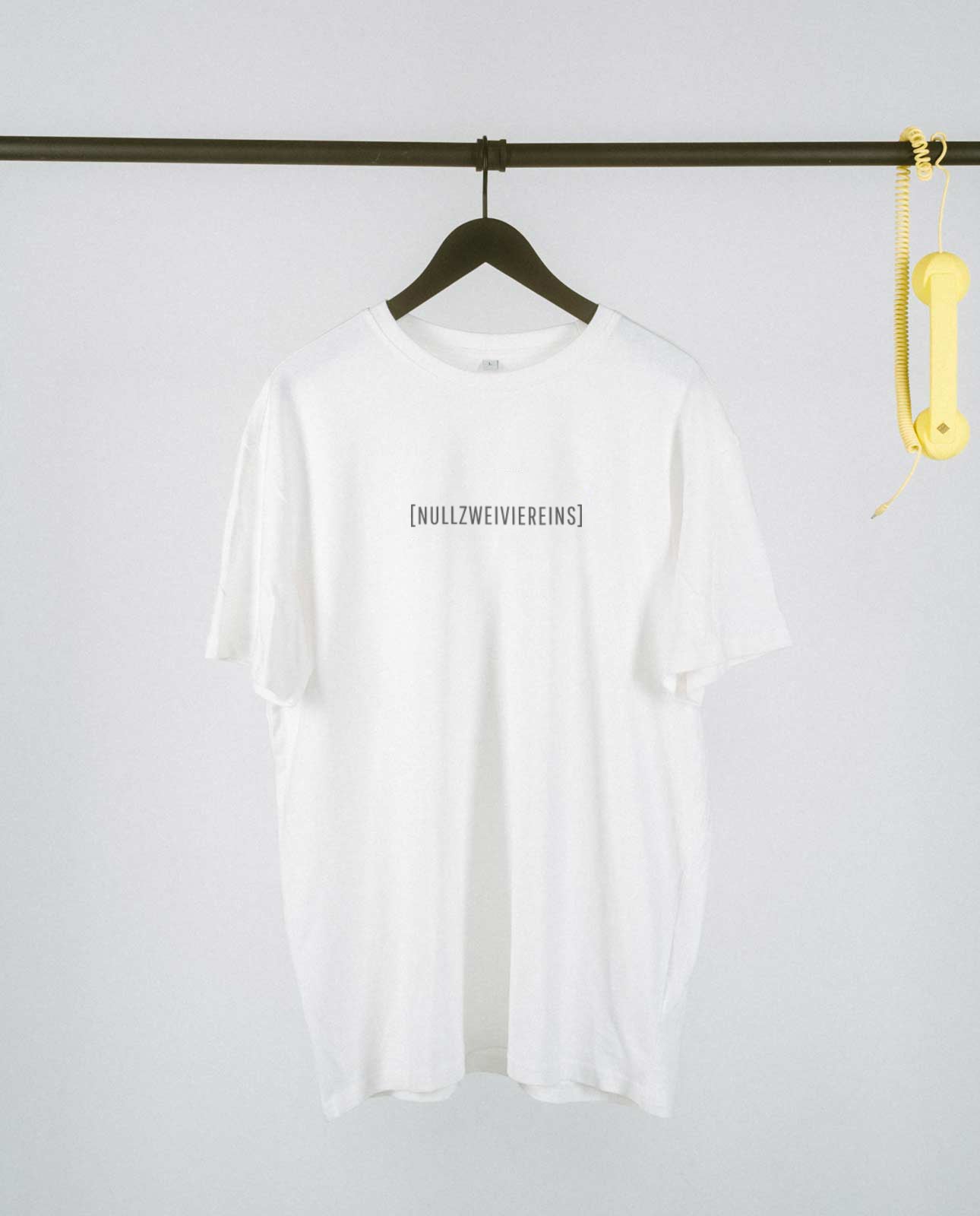 Weißes, locker geschnittenes T-Shirt, das auf einem schwarzen Kleiderbügel an einer schwarzen Metallstange hängt. Mittig auf der Brust steht in schlichter schwarzer Schrift „[NULLZWEIVIEREINS]“. Rechts an der Stange baumelt ein gelber Telefonhörer mit Spiralkabel als dekoratives Element. Der Hintergrund ist schlicht und hell, wodurch das T-Shirt im Vordergrund klar zur Geltung kommt.