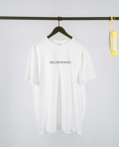 Weißes, locker geschnittenes T-Shirt, das auf einem schwarzen Kleiderbügel an einer schwarzen Metallstange hängt. Mittig auf der Brust steht in schlichter schwarzer Schrift „[NULLZWEIVIEREINS]“. Rechts an der Stange baumelt ein gelber Telefonhörer mit Spiralkabel als dekoratives Element. Der Hintergrund ist schlicht und hell, wodurch das T-Shirt im Vordergrund klar zur Geltung kommt.