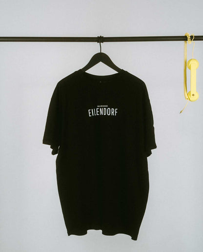 Schwarzes T-Shirt mit dem Schriftzug „EILENDORF“ in schlichten, weißen Großbuchstaben, mittig auf der Brust. Über dem Ortsnamen steht in kleinerer Schrift die Lautschrift [nʏlʦvaɪfɪaɪns], was dem Aachener Postleitzahlenbereich entspricht. Das minimalistische Design hebt den Stadtteil Eilendorf stilvoll hervor und sorgt für einen modernen, urbanen Look.