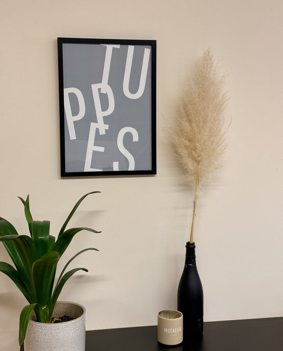 oster mit dem großformatigen Schriftzug „TUPPES“ in Weiß auf graublauem Hintergrund, gerahmt in Schwarz. Daneben Pampasgras in schwarzer Flasche, eine beige Tasse mit „MUCKELIG“-Aufdruck und eine grüne Pflanze im hellen Blumentopf.