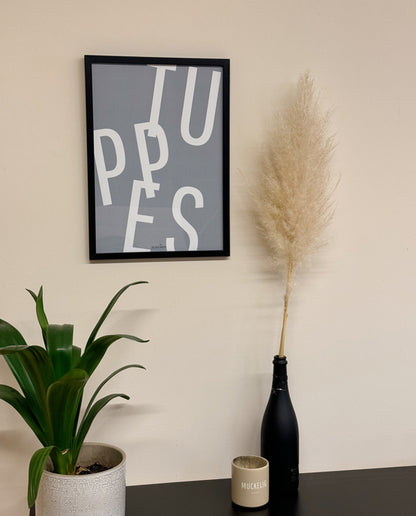 oster mit dem großformatigen Schriftzug „TUPPES“ in Weiß auf graublauem Hintergrund, gerahmt in Schwarz. Daneben Pampasgras in schwarzer Flasche, eine beige Tasse mit „MUCKELIG“-Aufdruck und eine grüne Pflanze im hellen Blumentopf.