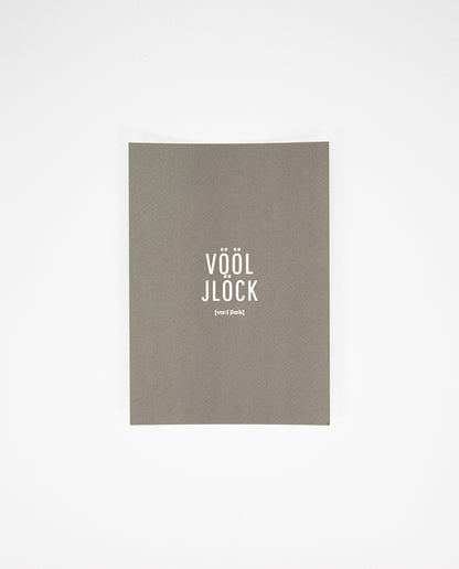 Graue Postkarte mit weißem Aufdruck in der Mitte: „VÖÖL JLÖCK“ in Großbuchstaben, darunter in kleinerer Schrift die Lautschrift.