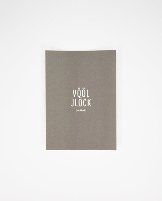 Graue Postkarte mit weißem Aufdruck in der Mitte: „VÖÖL JLÖCK“ in Großbuchstaben, darunter in kleinerer Schrift die Lautschrift.
