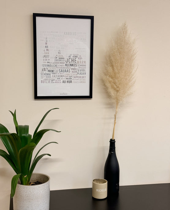 Poster mit einer Wortwolke aus Aachener Dialekt-Begriffen in Schwarz-Weiß in einem schwarzen Rahmen. Daneben Pampasgras in einer schwarzen Flasche, eine Zimmerpflanze im hellen Blumentopf und eine beige Tasse mit „MUCKELIG“-Aufdruck auf dunklem Tisch.