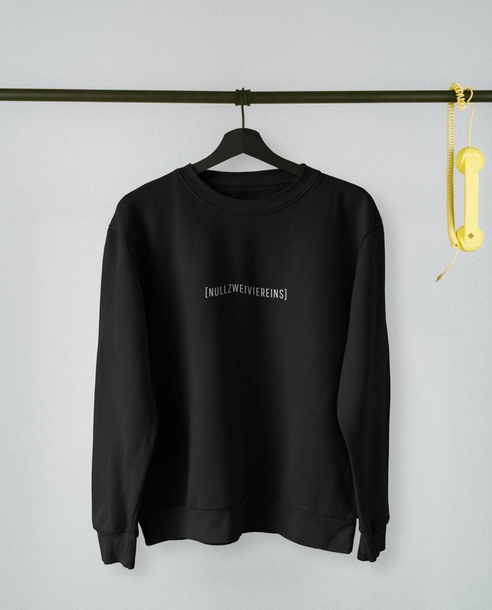 Schwarzes Sweatshirt mit weißem Schriftzug „[NULLZWEIVIEREINS]“ hängt auf einem schwarzen Kleiderbügel an einer Metallstange, rechts daneben ein gelber Telefonhörer. Hintergrund in hellem Grau.